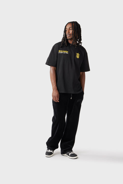 Front view of the model wearing the black t‑shirt with yellow chest logos. The small raised embroidered 'DL' monogram sits to the left chest and printed 'Dirty London' script sits on the right chest, while the crew neck and relaxed fit complement the look. The model pairs the t‑shirt with black track pants.