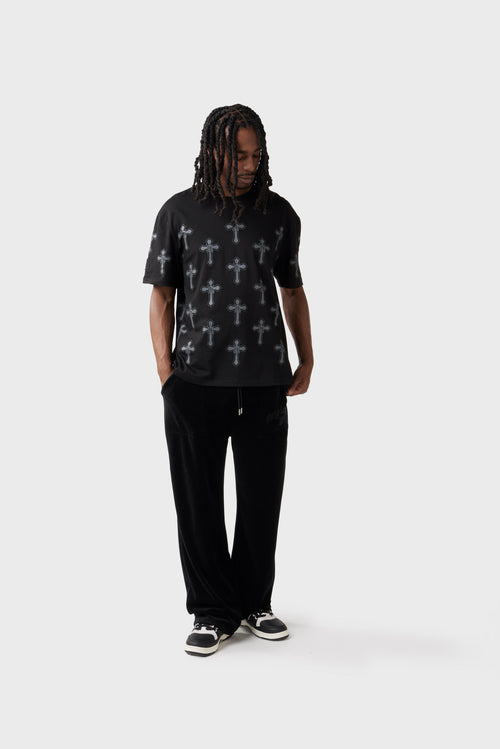 Model wearing black oversized T‑shirt covered in a repeating grey cross pattern with 'DL' monogram insert into each cross. The right sleeve carries a tonal embroidered 'DL' monogram. The shirt has a ribbed crew neck and relaxed fit, paired with black trousers.