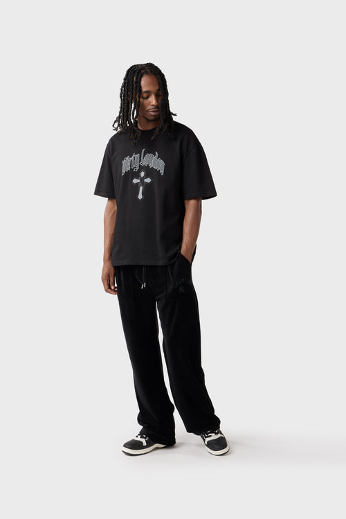 Front view of the model wearing the black t‑shirt featuring a grey cross with 'DL' monogram diamante and grey 'Dirty London' script across the chest. The relaxed cut and ribbed neckline create a casual look, and the model pairs the t‑shirt with black track pants.