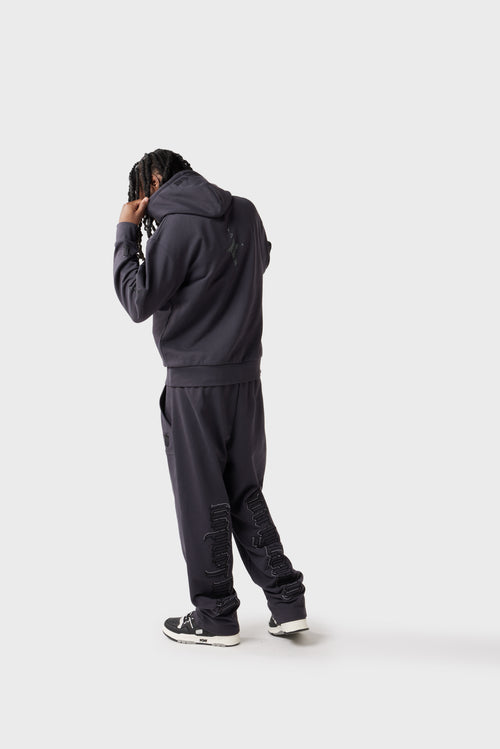 Back view of the model adjusting the hood while wearing the dark charcoal track pants. Vertical 'Dirty London' raised embroidery down both pant leg is clearly visible and a star motif appears on the back of the hoodie. The open cuffs enhance the relaxed vibe.