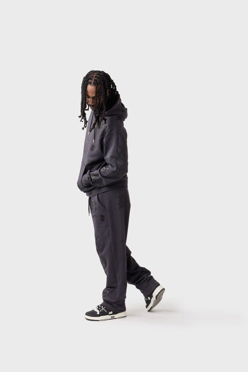 Front view of the model wearing dark charcoal open‑leg track pants with an all‑over tonal 'Dirty London' letter print. The pants feature an elasticated drawstring waistband with silver eyelets, front patch pockets and a black embroidered 'DL' monogram near the left hip.