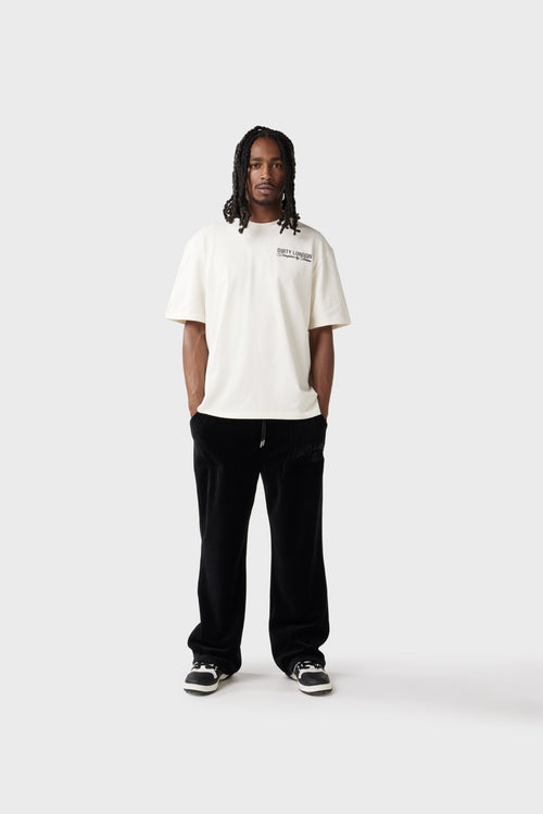 Front view of the model wearing a cream oversized T‑shirt with a black printed 'Dirty London' logo and cursive tagline 'Disruptive by Nature' embroidered on the left chest. The shirt has a relaxed fit and ribbed crew neck, paired with black track pants.