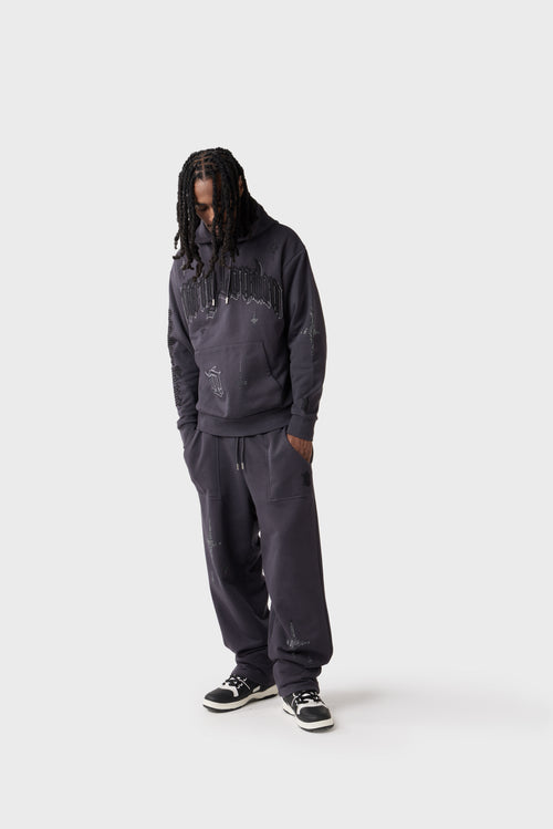 Full-body front view of model wearing dark charcoal oversized hoodie and matching track pants. The hoodie features a large embroidered 'Dirty London' logo across the chest, star motifs scattered across the chest and sleeves, 'DL' monogram on pocket and a drawstring hood. The matching pants have diamante stars on the front thigh and a vertical 'Dirty London' script down the back of the legs.