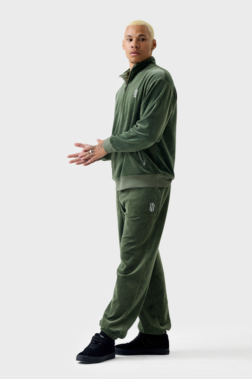 Full‑body view of a model wearing thyme‑green velour cuffed track pants with a matching track jacket. The pants have a drawstring waistband, rhinestone 'DL' patch pocket, side pockets, and cuffed ankles.