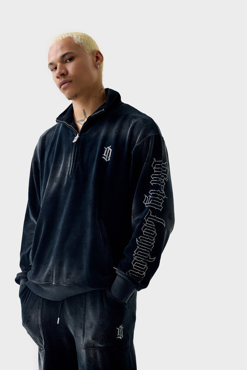 Front view of a model wearing a black acid‑wash velour half‑zip track jacket with all over debossed 'DL' repeat pattern; rhinestone 'DL' monogram on chest, rhinestone Dirty London script down left sleeve, side zip pockets and ribbed cuffs and hem.