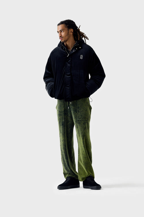 Full‑body front view of a model wearing cropped length black velour puffer jacket zipped up with high collar, embroidered diamanté ''DL'' monogram on chest and vertical repeat monogram pattern on sleeve, paired with green acid wash track bottoms.