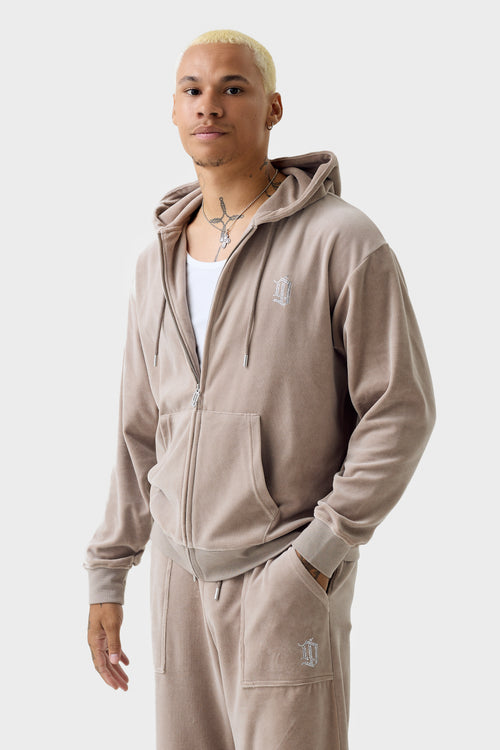 Front view of a model wearing a beige velour zip hoodie zipped halfway with matching pants; features hood with drawcords, kangaroo pocket, rhinestone ''DL'' monogram on chest, and ribbed cuffs and hem.