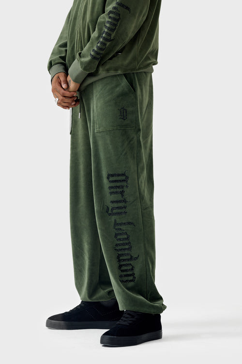 Side view of thyme green velour cuffed jogger on model wearing matching hoodie; shows black rhinestone Dirty London script down leg, patch pocket with monogram, drawstring waistband and elastic cuffs.