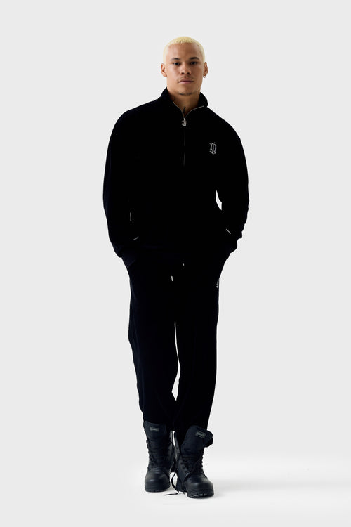 Full‑body view of model wearing black velour half‑zip track top and matching pants; top has a funnel neck with half zipper, diamanté ''DL'' monogram on chest, ribbed cuffs and hem and front pockets.