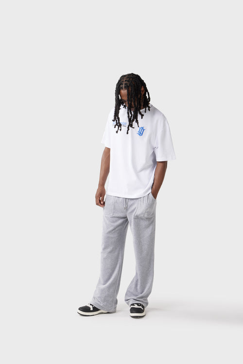 Front view of the model wearing the white t‑shirt with blue chest logos. The embroidered 'DL' monogram and printed 'Dirty London' script stand out against the white fabric, and the relaxed fit pairs with grey track pants.