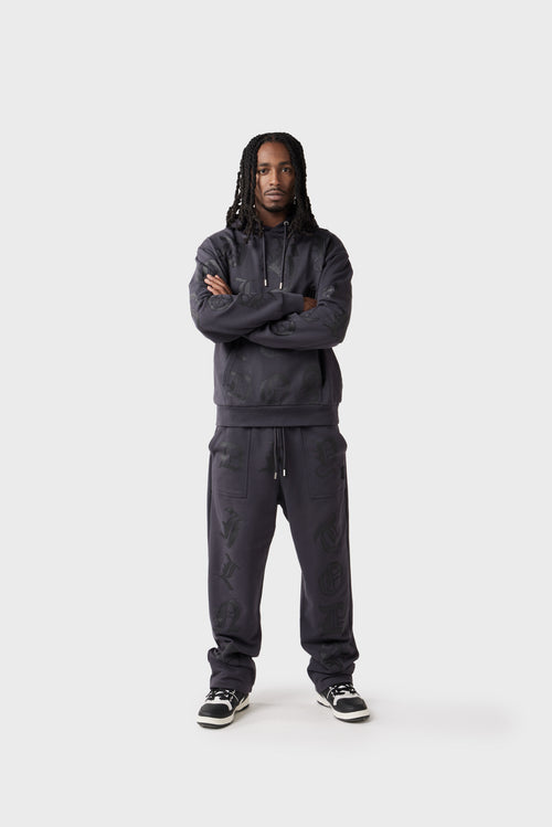 Front view of the model with arms crossed wearing a dark charcoal oversized hoodie. The hoodie features an all‑over tonal 'Dirty London' letter print with embroidered outline and a black raised embroidery 'DL' monogram on the left chest. A drawstring hood with silver metal hardware and a kangaroo pocket complete the relaxed design.
