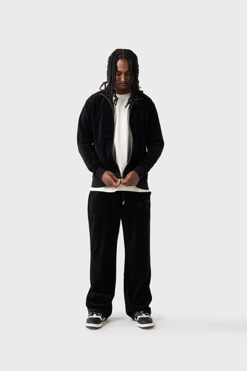 Front view of the model wearing the black velour track jacket zipped up. The diamante 'Dirty London' logo glistens on the chest, the high collar adds a sporty edge and zipped side pockets blend into the plush fabric. Paired with matching track pants for a sleek set.
