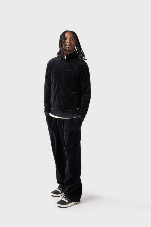 Front view of the model wearing the black velour track jacket zipped up. The diamante 'Dirty London' logo glistens on the chest, the high collar adds a sporty edge and zipped side pockets blend into the plush fabric. Paired with matching track pants for a sleek set.