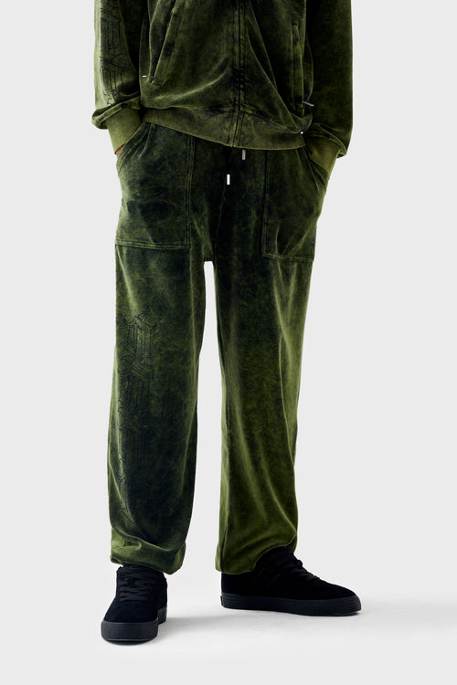 Full-body view of green acid-wash velour cuffed track pants worn by a model; drawstring waistband, patch pockets, rhinestone 'DL' repeat pattern running vertically down the right leg and elasticated cuffs.
