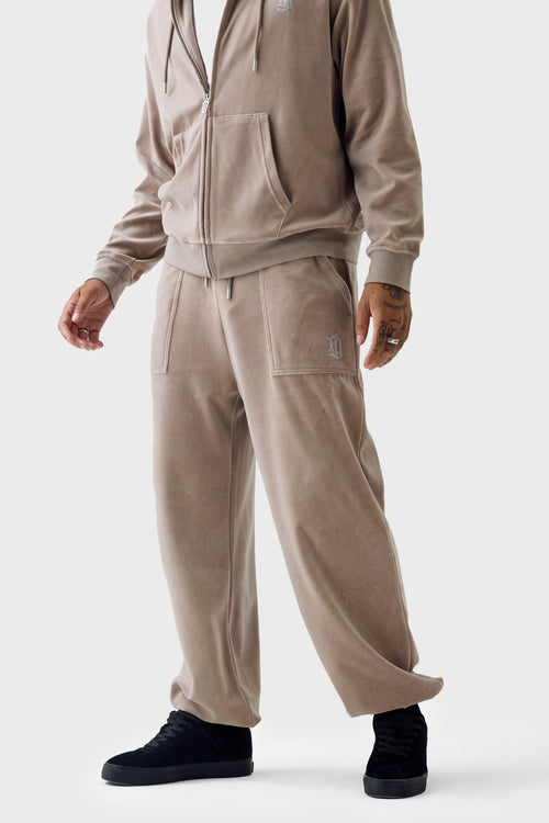 Full‑body view of the beige velour cuffed track pants featuring a drawstring waistband, rhinestone 'DL' patch pocket on the hip, side pockets, and elastic cuffs; relaxed fit and velour texture are visible.