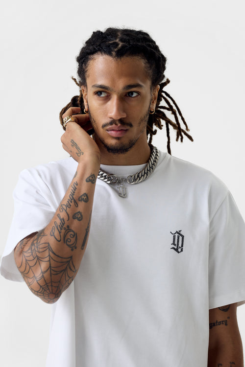 Front close-up of white T-shirt on model showing small rhinestone 'DL' monogram on left chest, crew neckline and oversized fit.