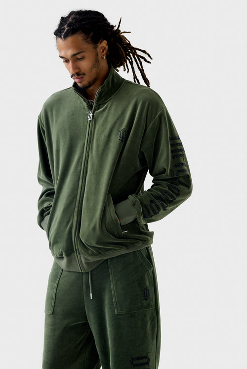 Front view of model wearing thyme green velour zip-through track jacket zipped up, featuring black rhinestone Dirty London script down left sleeve and diamanté 'DL' monogram on chest.