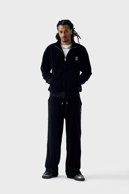 Full-body view of black velour straight track pants and matching jacket; pants have drawstring waistband, rhinestone 'DL' patch pocket on the thigh, relaxed wide leg and open ankles.