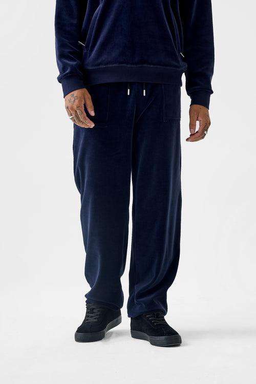Full‑body front view of the navy velour cuffed track pants featuring a drawstring waistband, rhinestone 'DL' patch pocket, side pockets, and elastic cuffs; the relaxed fit is evident.